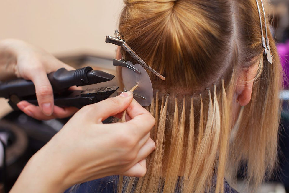Hair extensions applied to blonde hair using fusion technique. Hands and tools are visible. Hair clips secure sections. Salon setting.