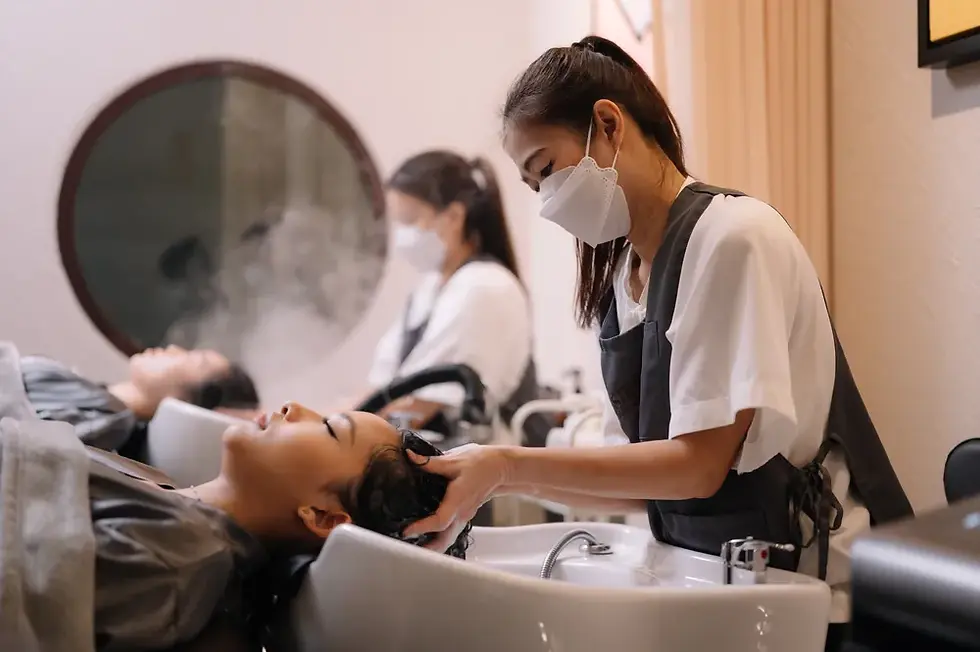 A stylist in a mask washes a client's hair at a salon with chairs and a round mirror. The client appears relaxed. Soft, neutral tones.
