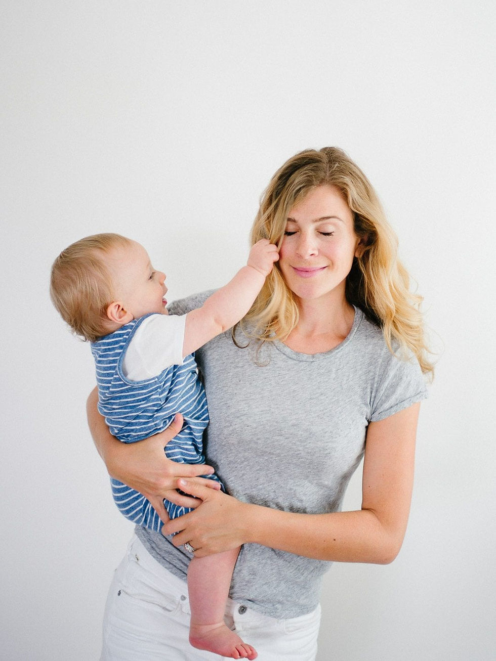 Woman holding a baby in a blue-striped romper. Baby playfully touches her hair. Both are smiling. White background, casual mood.