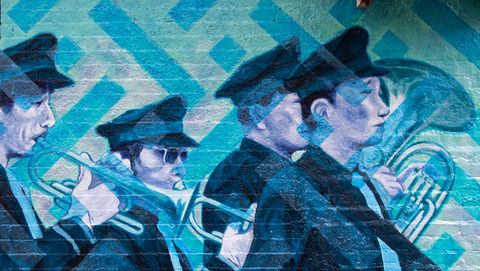 Chinese funereal marching bands are meant to ward off evil spirits and safeguard the recently deceased’s journey to the afterlife.  It is the living’s responsibility to shepherd their dead safely to the next phase, ensuring their spirits are provided for, loved, and avoid the fate of the hungry ghost: left to wander eternally, never reaching the next rung of reincarnation, forgotten by their families. 

The players often arrive by bus on Pike Street and East Broadway, the Buddhist temple in a former synagogue serving as a meeting place, site of the former and much beloved “99 Cent New BJ” everything shop long collectively mourned.  They tune up, mingle, and then march: up around East Broadway, past the future ghost office tower that glows purple and red at night, towards Boe Fook Funeral parlor on Canal St.  The cacophony ends by noon, a moment when other neighborhood fixtures typically only seen after sundown start to rouse.  If at night the stretch of Canal Street encompassing Boe Fook teems with the libidinal energy of a New York dream, its prosaic beauty shines democratically during the day, as locals conduct business and life amid the crooked streets.  

New York neighborhoods are live palimpsests: immigrant groups supplant each other, different cultures take cues from each other, populations coexist, in both harmony and tension.  Our micro-neighborhood now half-jokingly known as “Dimes Square,” a sleepy corner of Lower Manhattan and destination on the road to nowhere, is enjoying a moment of cultural shine, a beacon of white hot nonchalance swirling amidst an entrenched Chinese immigrant population itself living among the signifiers of earlier Jewish and Italian communities.  The funereal marching bands themselves are a cultural mashup, borne out of Buddhist tradition, fused with Western-style military rites in British colonial Hong Kong, and more locally, historically composed of Italian-American neighbors amid the Chinese mourners.

Jakob Bokulich’s Every Day in Dimes Square Begins with a Funeral, a mural on that tiny stretch of Orchard below Canal St., depicts a funeral marching band behind a flower laden casket, coolly recast in blues and greens. The figures are larger than life, the mural modest in scale, sandwiched between the perennially-packed Kiki’s and a smoke shop.  The permanent visual presence of the marchers rendered in a style miming socialist realism, lending gravity and steely optimism, serves as a marker of a neighborhood in constant flux.  By memorializing the memorializers, Bokulich extends the celebratory cycle of life, death, and rebirth to the entire neighborhood.  The ghosts of long-lost denizens of the demi-monde, dead or moved on, abut those of Chinese neighborhood elders, every single one displaced in some way, every single one deserving an auditory ovation, a boisterous hello and good-bye, a cacophonous warning to the morass of evil spirits potentially lurking around every corner.

David Fierman, Fierman Gallery