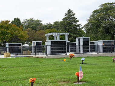 Fenced columbarium garden overlooking cemetery lawn with flowers and an American flag.