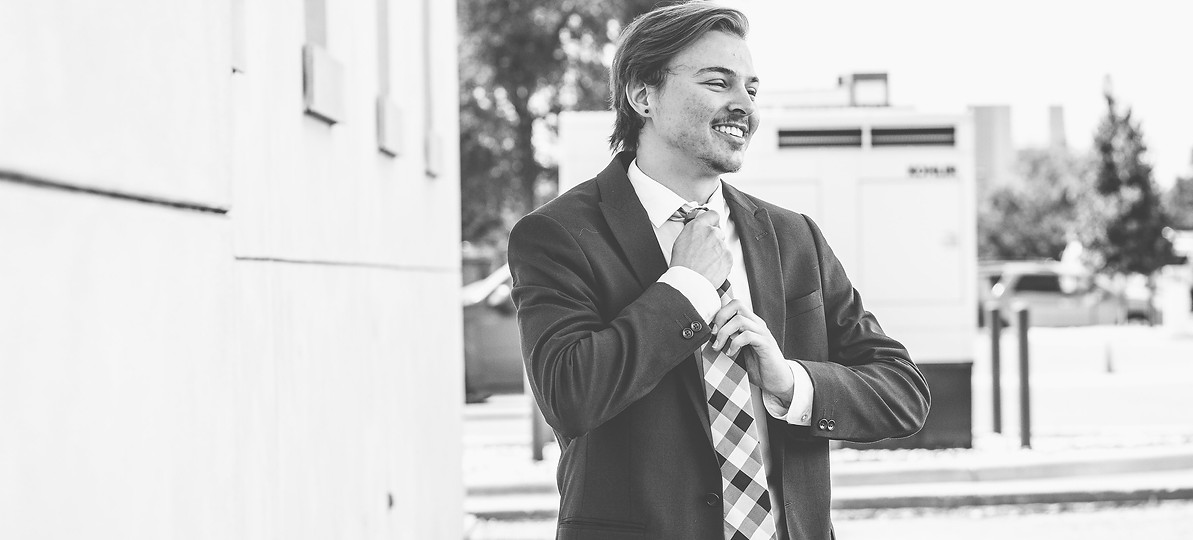 A black and white photo of a masculine person looking away from the camera while adjusting his tie.