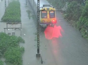 तेज़ बारिश में पानी से भरे ट्रैक से गुज़रती ट्रेन, पानी पर लाल टेल लाइट की चमक और सड़क के किनारे हरी-भरी हरियाली।