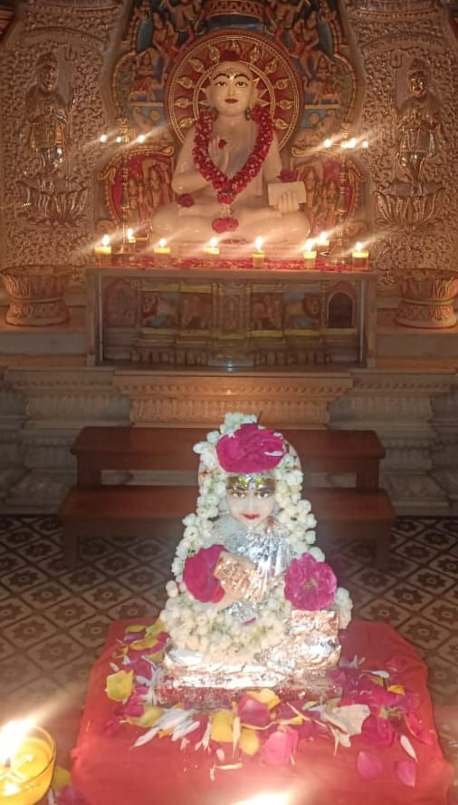 Temple shrine with two adorned idols, lit oil lamps, flowers, and carved stone backdrop.