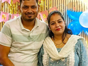 एक मुस्कुराता हुआ जोड़ा किसी पार्टी में पोज़ दे रहा है, जिसके बैकग्राउंड में सुनहरे स्ट्रीमर, गुलाबी बंटिंग और नीले गुब्बारे लगे हैं।