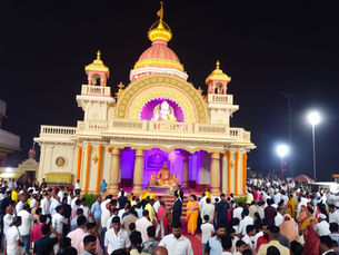 रात के समय एक शानदार, सुनहरी रोशनी वाले मंदिर में भीड़ जमा है; वहाँ बैंगनी रोशनी के नीचे एक चमकती हुई मूर्ति है और उसके पीछे अंधेरा आसमान है।