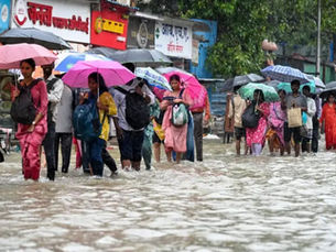 बारिश और अफरा-तफरी वाले माहौल में, लोग रंग-बिरंगी छतरियां लिए पानी से भरी सड़क पर चल रहे हैं और हिंदी साइनबोर्ड वाली दुकानों के पास से गुज़र रहे हैं।