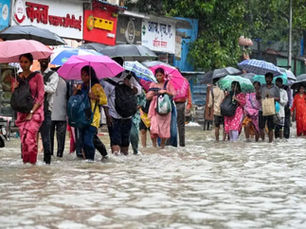 बारिश और अफरा-तफरी वाले माहौल में, लोग रंग-बिरंगी छतरियां लिए पानी से भरी सड़क पर चल रहे हैं और हिंदी साइनबोर्ड वाली दुकानों के पास से गुज़र रहे हैं।