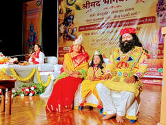 खास वेशभूषा पहने कलाकार हिंदी बैनर के सामने मंच पर बैठे हैं; बाईं ओर एक स्पीकर रखा है और माहौल भक्तिपूर्ण व उत्सव जैसा है।