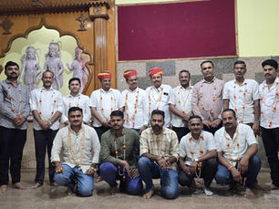 सफ़ेद शर्ट पहने और गले में माला डाले पुरुषों का एक समूह, एक औपचारिक जश्न के मौके पर मूर्तियों वाले हिंदू मंदिर के सामने पोज़ दे रहा है।