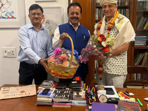 कार्यालय में तीन पुरुष मुस्कुराते हुए फल-टोकरा और फूलों का गुलदस्ता पकड़े हैं; मेज पर किताबें व स्टेशनरी हैं।