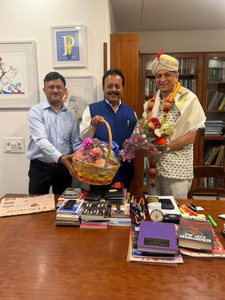 कार्यालय में तीन पुरुष मुस्कुराते हुए फल-टोकरा और फूलों का गुलदस्ता पकड़े हैं; मेज पर किताबें व स्टेशनरी हैं।