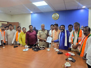 एक कॉन्फ्रेंस रूम में, BJP के स्कार्फ़ पहने और नीले बैकड्रॉप के सामने, एक पुलिस अफ़सर और नेता टेबल के चारों ओर कागज़ों का आदान-प्रदान कर रहे हैं।