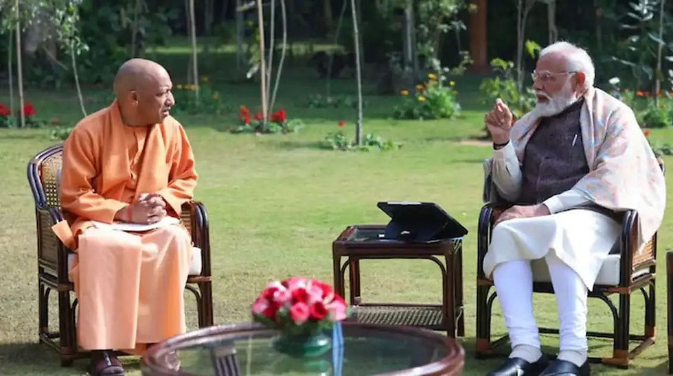 दो आदमी एक बगीचे में बैठकर बातें कर रहे हैं; एक ने केसरिया कपड़े पहने हैं और दूसरे ने सफ़ेद। मेज़ पर एक टैबलेट रखा है और सामने फूल हैं।