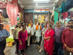 एक छोटी सी कपड़ों की दुकान में कुछ वयस्क पोज़ दे रहे हैं; ऊपर रंग-बिरंगे कपड़े लटके हुए हैं और एक साइनबोर्ड पर 'H Halluram TRADERS' लिखा है।