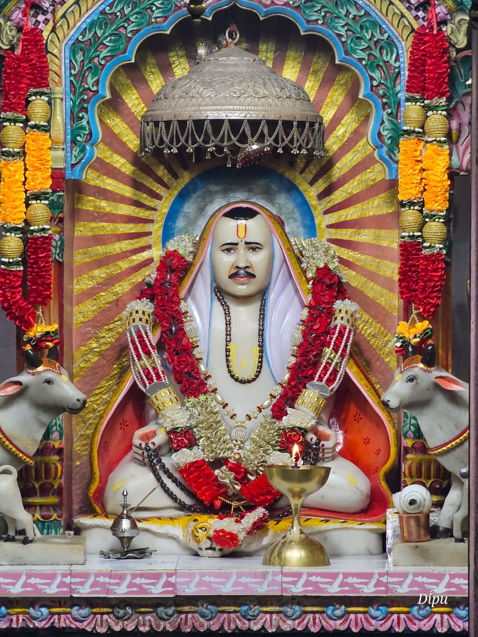 Hindu shrine with a garlanded seated deity, lamps, and cow statues under a colorful arch; Dipu watermark.