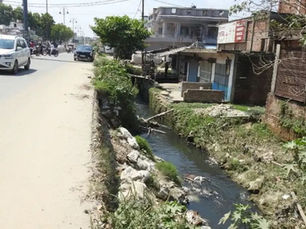 एक खुली जल निकासी नहर के बगल में एक धूपदार शहरी सड़क पर सफेद एसयूवी, ईंट की इमारतों और पास में कुछ दूर के वाहनों के साथ।