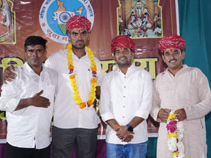 सफ़ेद और हल्के भूरे रंग की शर्ट और लाल पगड़ी पहने चार आदमी एक मंच पर पोज़ दे रहे हैं; उनमें से एक ने हिंदी बैनर के सामने गेंदे के फूलों की माला पहनी हुई है।
