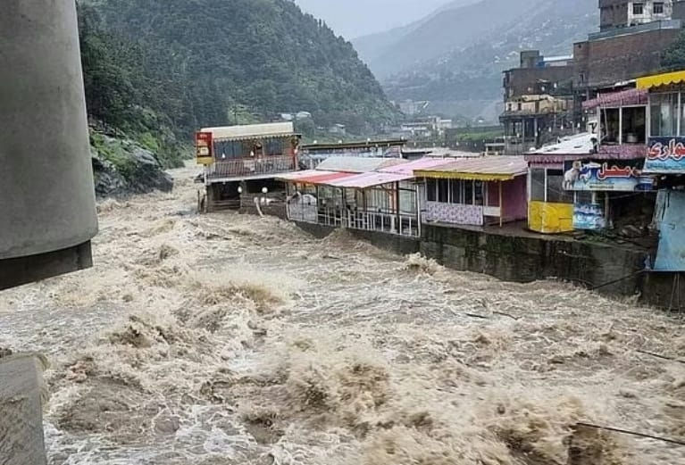 बादलों से घिरी पहाड़ियों और मटमैले आसमान के नीचे, अरबी साइनबोर्ड वाली दुकानों के बगल से पहाड़ी कस्बे में बाढ़ का उग्र पानी तेज़ी से बह रहा है।