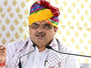 सुनहरे पैटर्न वाले बैकग्राउंड के सामने, रंग-बिरंगी पगड़ी पहने, सफ़ेद शर्ट और पैटर्न वाला स्कार्फ़ ओढ़े एक व्यक्ति माइक्रोफ़ोन पर बोल रहा है।