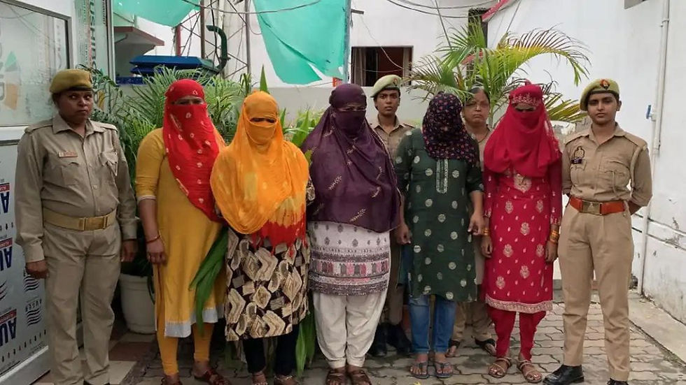 एक आंगन में पुलिस अधिकारियों के साथ घूंघट किए हुए छह महिलाएं खड़ी हैं, सफेद दीवारों और हरियाली के सामने रंग-बिरंगी पोशाकें और वर्दी पहने हुए, गंभीर मुद्रा में