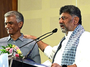 घर के अंदर एक पोडियम पर दो आदमी हैं; एक आदमी दूसरे के कंधे पर हाथ रखकर बात कर रहा है; सामने माइक्रोफ़ोन और फूल रखे हैं, और माहौल खुशनुमा है।