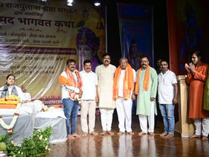 एक हिंदू सांस्कृतिक कार्यक्रम में कुर्ता और शर्ट पहने पुरुष मंच पर पोज़ दे रहे हैं; साथ ही, एक महिला सजी-धजी मेज़ के पास है और पीछे हिंदी में लिखा बैनर लगा है।