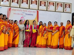एक हॉल में, नारंगी-पीली साड़ियाँ पहने महिलाएँ गर्व और उत्सव के माहौल में, फ़्रेम की हुई तस्वीरों के नीचे हाथ में तोहफ़े लिए हिंदी कार्यक्रम के बैनर के सामने पोज़ दे रही हैं।