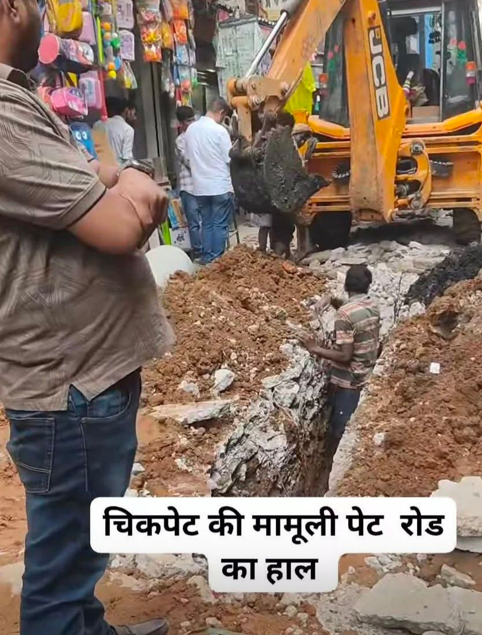 Excavator lifts rubble from a collapsed road in a busy market street, with onlookers nearby and Hindi text overlaid.