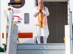 विमान की सीढ़ियों पर हाथ जोड़कर खड़े नरेंद्र मोदी, सफेद-गुलाबी पोशाक व केसरिया दुपट्टा, पीछे विमान दरवाज़ा खुला है।