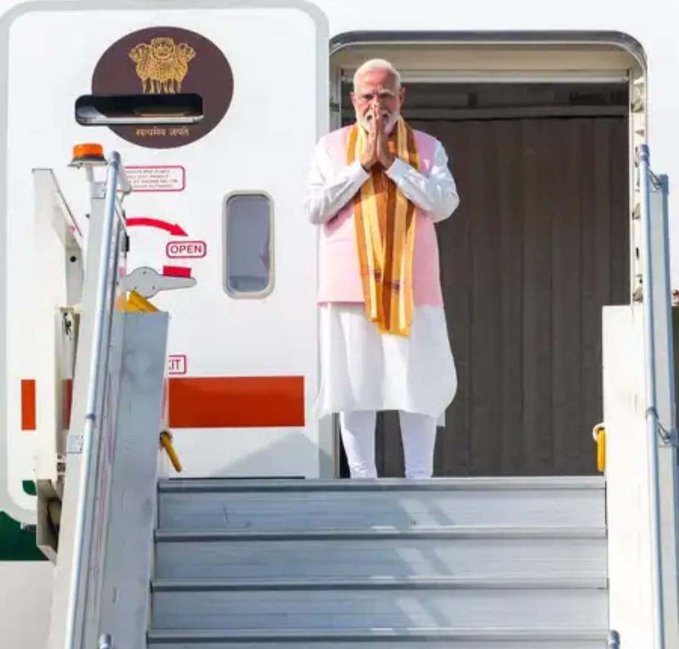 विमान की सीढ़ियों पर हाथ जोड़कर खड़े नरेंद्र मोदी, सफेद-गुलाबी पोशाक व केसरिया दुपट्टा, पीछे विमान दरवाज़ा खुला है।