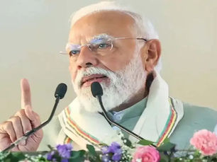 नरेंद्र मोदी एक सादे हल्के रंग के बैकग्राउंड के सामने पोडियम पर खड़े होकर बोल रहे हैं; उनके सामने माइक और फूल रखे हैं और वे अपनी उंगली उठाए हुए हैं।