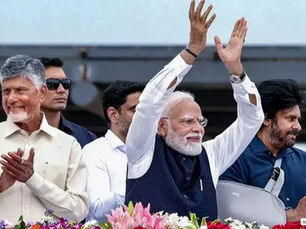 Narendra Modi waves both hands at a public event as men applaud beside him, with flowers in front and an upbeat mood.
