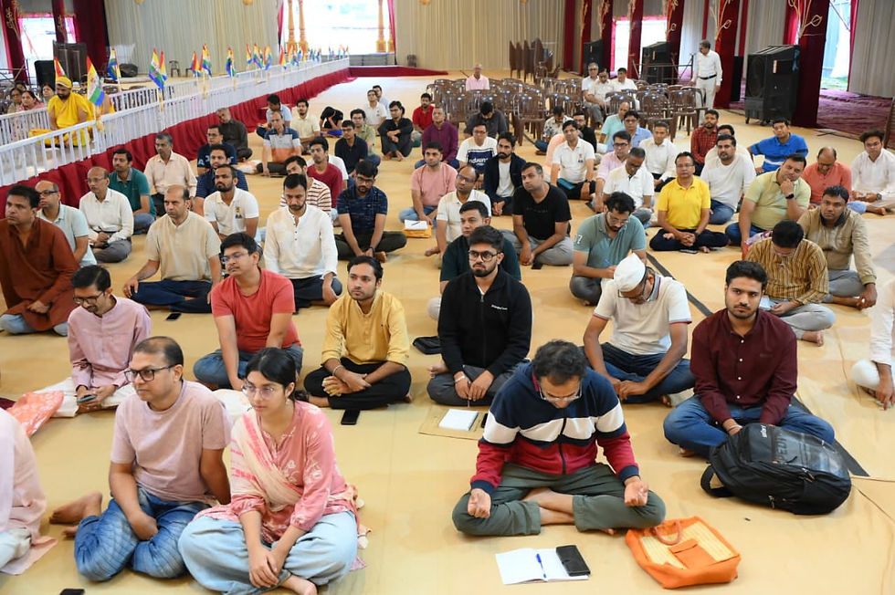 हॉल में कई लोग फर्श पर बैठे ध्यान/प्रार्थना कर रहे हैं; पीछे छोटी रंग-बिरंगी झंडियाँ, कुर्सियाँ और शांत सभा माहौल।