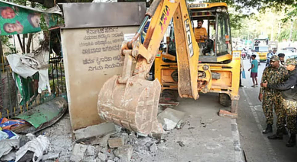 एक व्यस्त सड़क पर सैनिक देखते हुए, एक पीला बैकहो सड़क के किनारे बनी दीवार को गिरा रहा है।