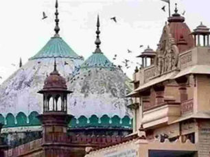 हल्के रंग के आसमान के सामने, नक्काशीदार मंदिर के गुंबदों और हल्के भूरे रंग के टॉवर के ऊपर पक्षी उड़ रहे हैं, जिससे एक शांत और ऐतिहासिक नज़ारा बन रहा है।
