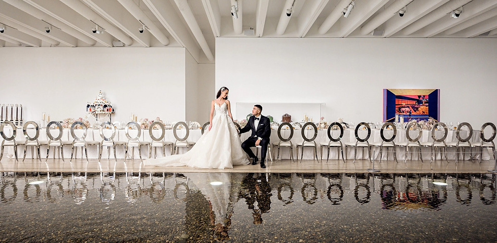 Bride in white gown and groom in black tuxedo pose at a long wedding table in a bright modern hall beside a reflective pool