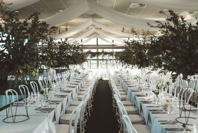 Elegant wedding reception setup with long white-draped tables, white chairs, greenery, and draped ceiling in a bright hall.