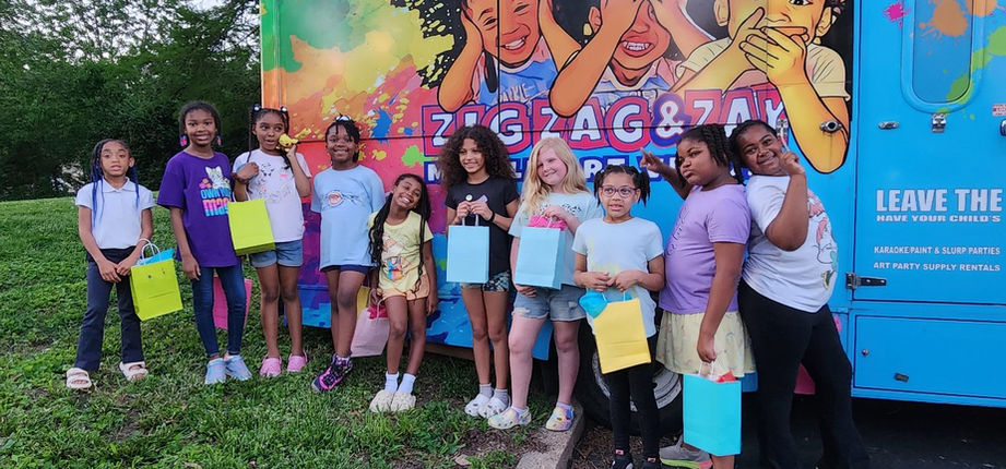 Happy diverse girls holding gift boxes beside ZIG ZAG & ZAY bus.