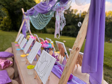 Outdoor art party setup with easels, canvases, purple decor for children.