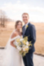 Bride and groom smiling, holding yellow and white bouquet. She wears a flowing dress; he a navy suit. Bright, open field background.