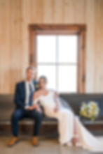 Bride and groom sit together on a bench in front of a window. She wears a white dress with a bouquet beside her; he wears a navy suit.