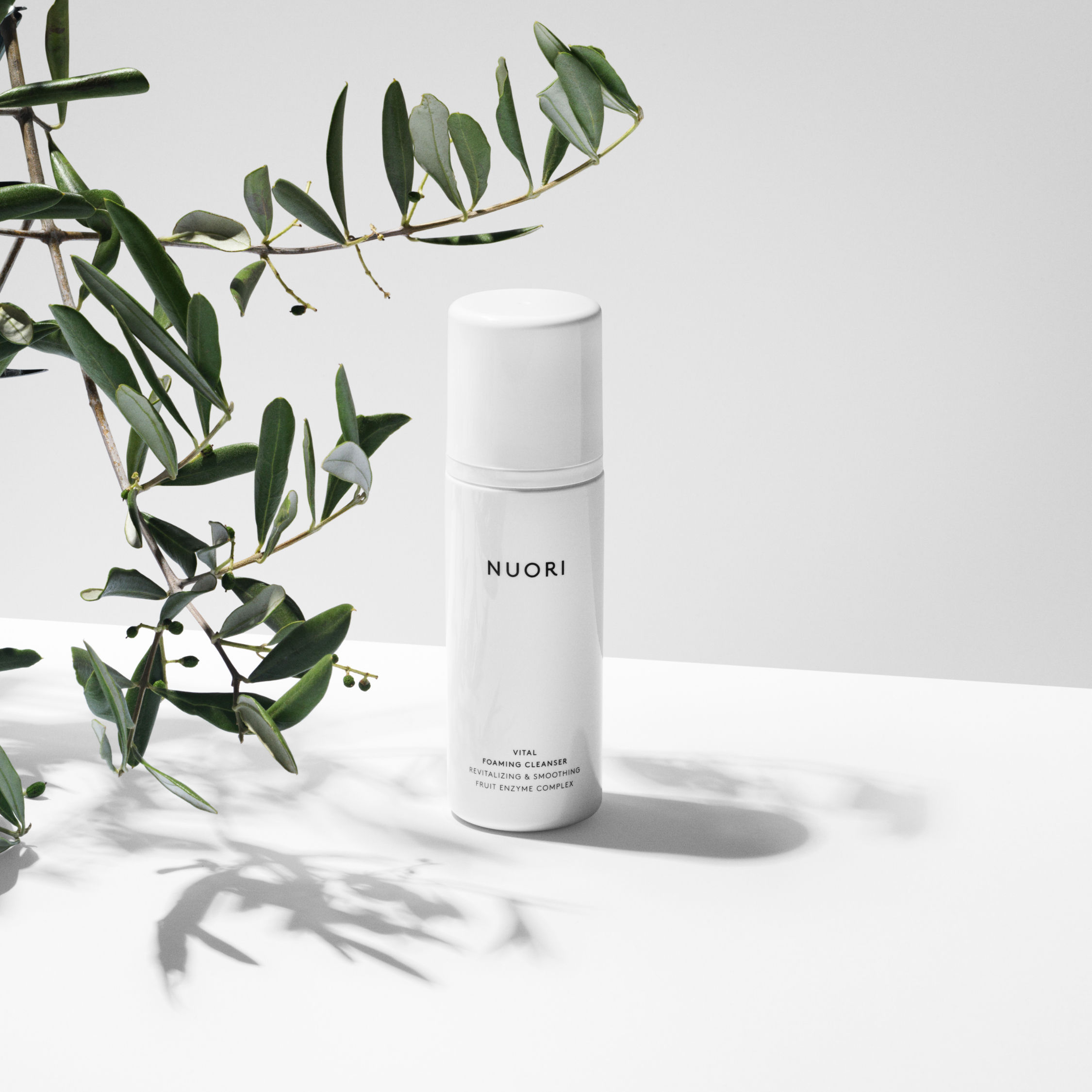 NUORI Vital Foaming Cleanser u minimalističkoj beloj bočici pored maslinove grane na svetloj podlozi.