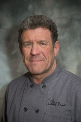 Portrait of Chef Brad Petersen wearing a gray double-breasted chef’s coat embroidered with his name. He has short, curly brown hair and a confident expression. The background is a softly lit, textured gray backdrop, giving a professional and refined look.