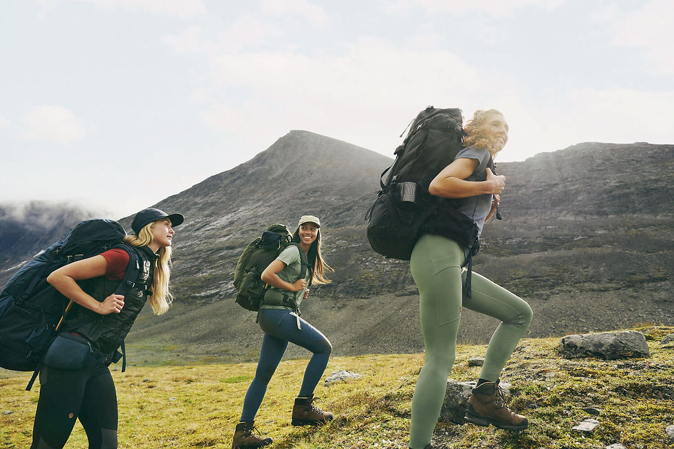 Three smiling women hike uphill with large backpacks in a rocky mountain landscape under a bright cloudy sky.