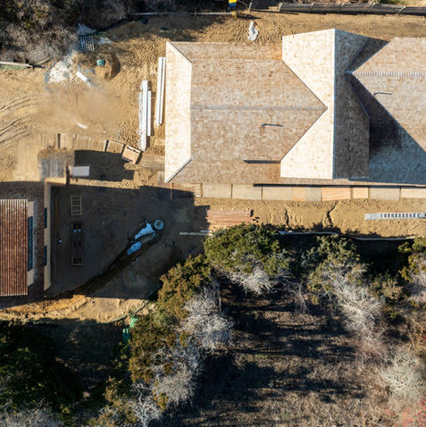 Top-down aerial view of an in-progress coastal home construction site highlighting roof layouts, connected structures, and surrounding landscape