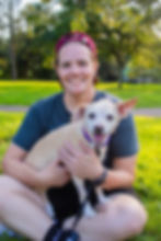 A portrait of Meghan Pellerin and Reilly, a small blonde dog with short hair and one floppy ear. They are in a park. They are looking at the camera and smiling