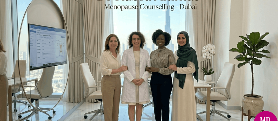 A group of four smiling women standing in a modern, professional office in Dubai with floor-to-ceiling windows. From left to right: a woman in a light tan shirt and pants, Dr. Kubra Altintas in a white doctor's lab coat, a woman in a patterned blouse and black pants, and a woman in a long white dress and green hijab. They are linked arm-in-arm. The background window shows a clear view of the Dubai skyline, including the Burj Khalifa. A large monitor on the left shows medical information. Above the women, text reads "Dr Kubra Altintas - Menopause Counselling - Dubai". A pink logo in the bottom right corner says "MD KUBRA ALTINTAS".