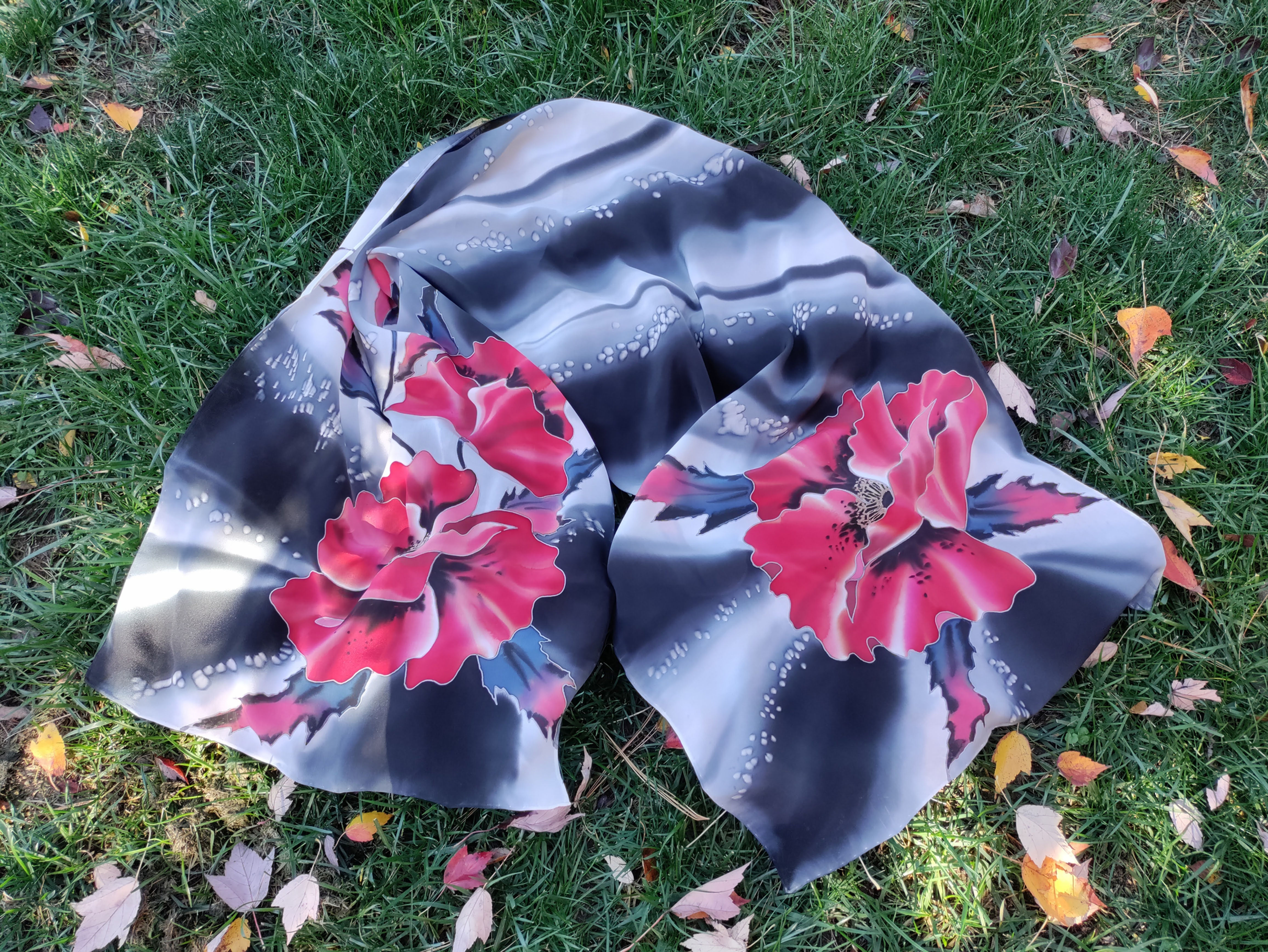 Hand-painted silk scarf with red poppies laid out flat on grass, showing full floral pattern on a gray-black background.