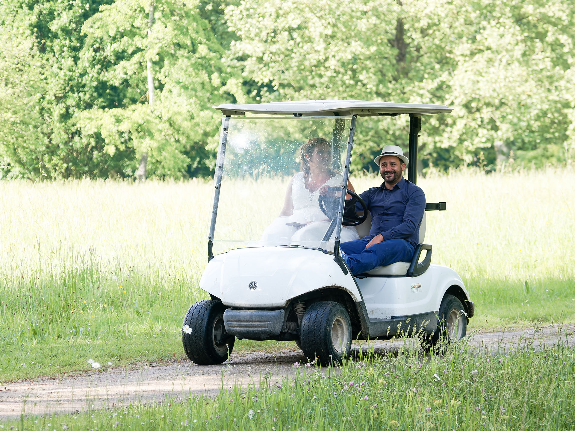 Photo de mariage prise au Domaine de Courson en Essonne, montrant la mariée Audrey conduisant une voiturette de golf à travers les allées fleuries du parc, accompagnée d’un invité complice. Ce moment de respiration et de spontanéité s’inscrit dans le cadre verdoyant et champêtre du lieu de réception. Image capturée sur le vif par une photographe professionnelle spécialisée dans les reportages de mariage naturels en Essonne et région parisienne.